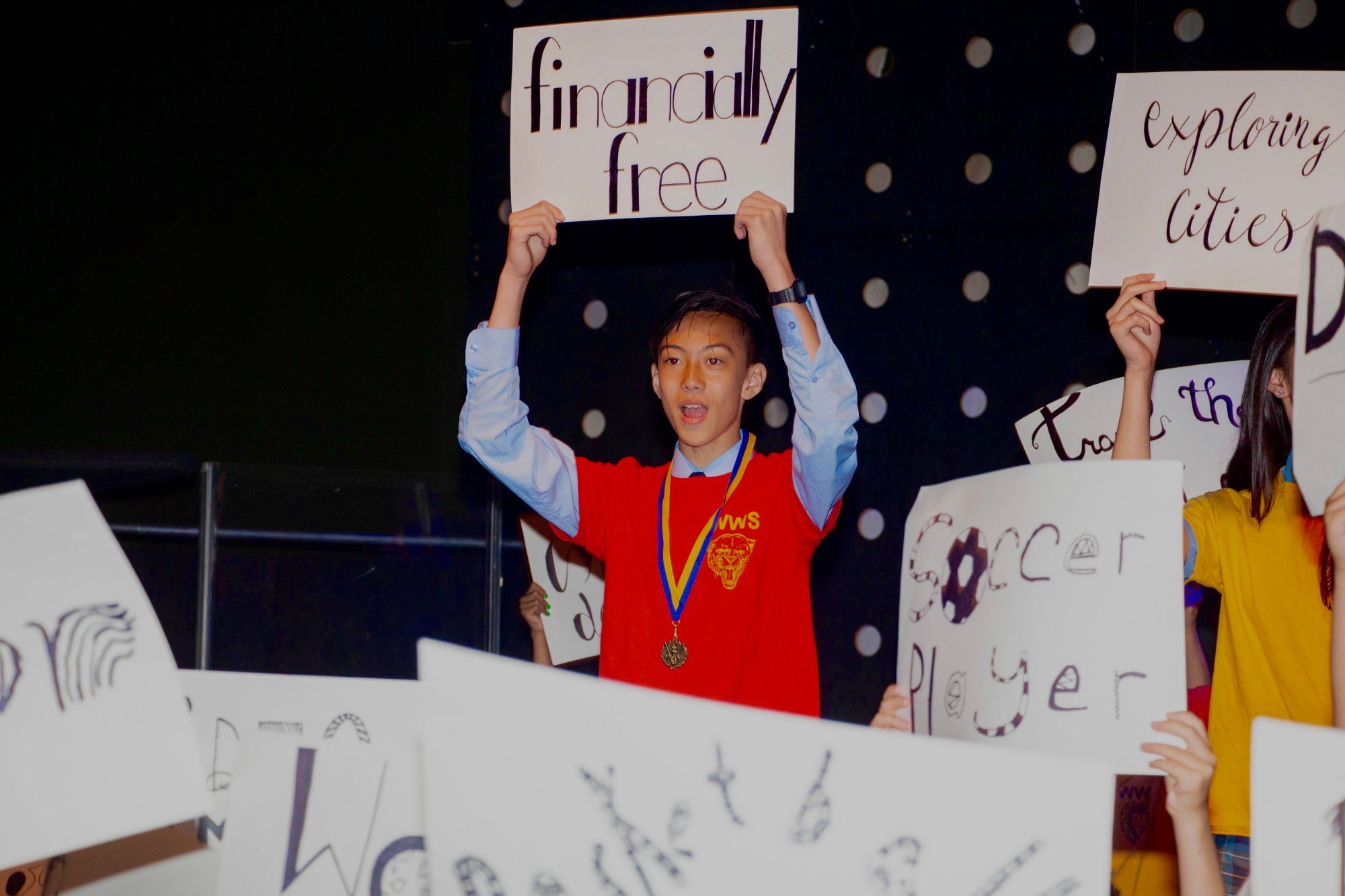 Ethan at his Grade 8 graduation in 2019, holding up a sign that reads “financially free” among classmates’ signs reading “exploring cities”, “soccer player” and others